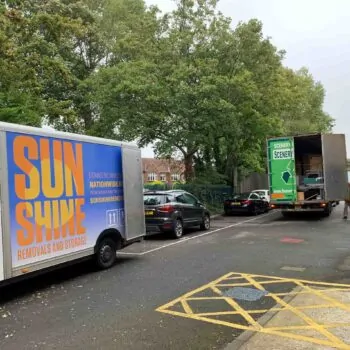 Two Sunshine Removals moving vans are parked on a street lined with trees. A man observes the scene.