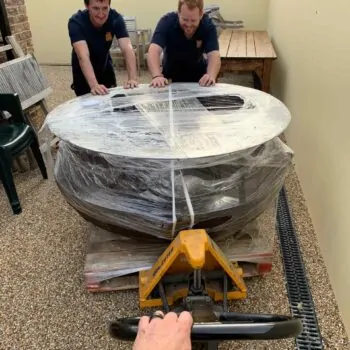 Two men in blue shirts push a large, wrapped table on a pallet truck. A third man pulls the pallet truck handle.