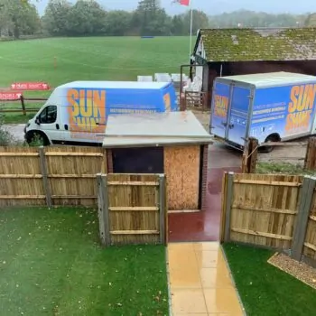 Two Sunshine Removals vans parked near a wooden fence. A large green sports field and an older building with a mossy roof are in the background.